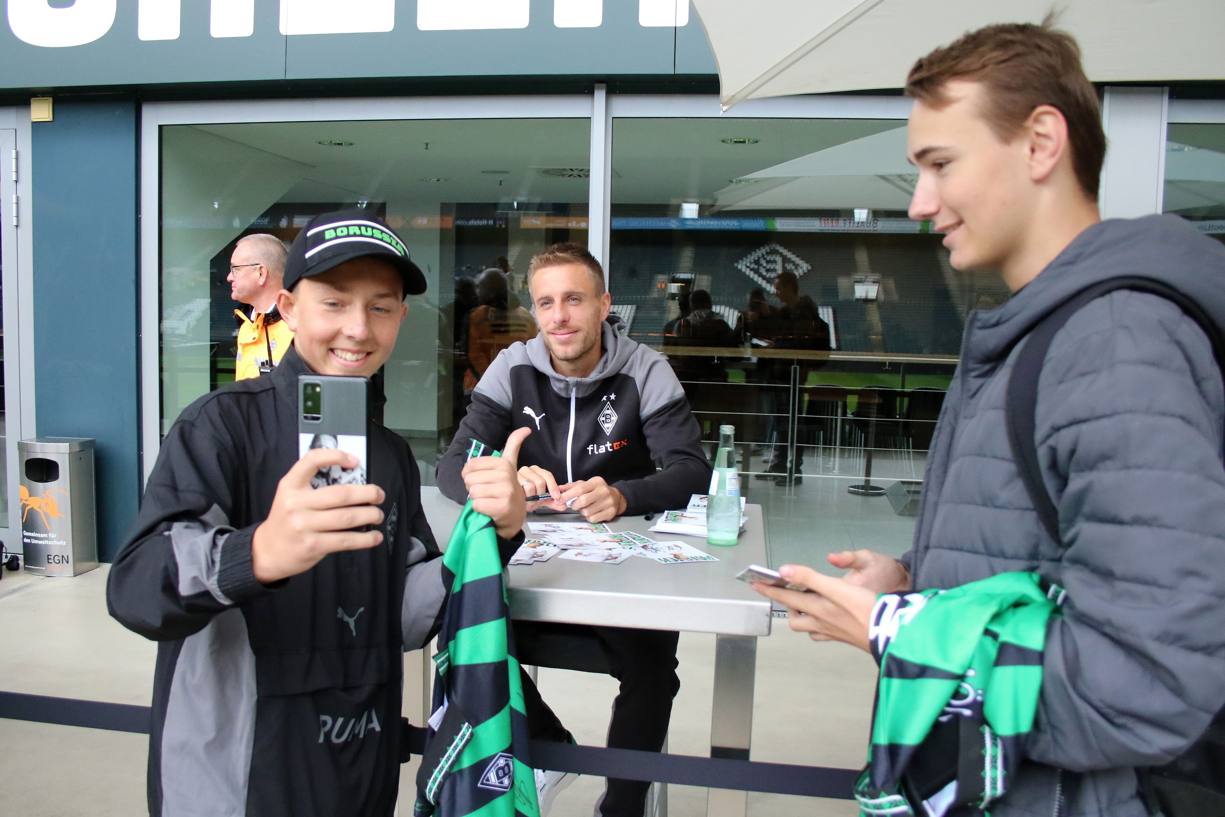 Herr Herrmann, ein Selfie bitte! Die Gladbach-Profis wie Patrick Herrmann haben sich zur Saisoneröffnung am Sonntag (6. August 2023) den Fans hautnah für Autogrammwünsche gestellt. Familienfest bei Borussia. Herrmann lächelt und sitzt an einem Tisch, derweil machen Fans gemeinsame Fotos.