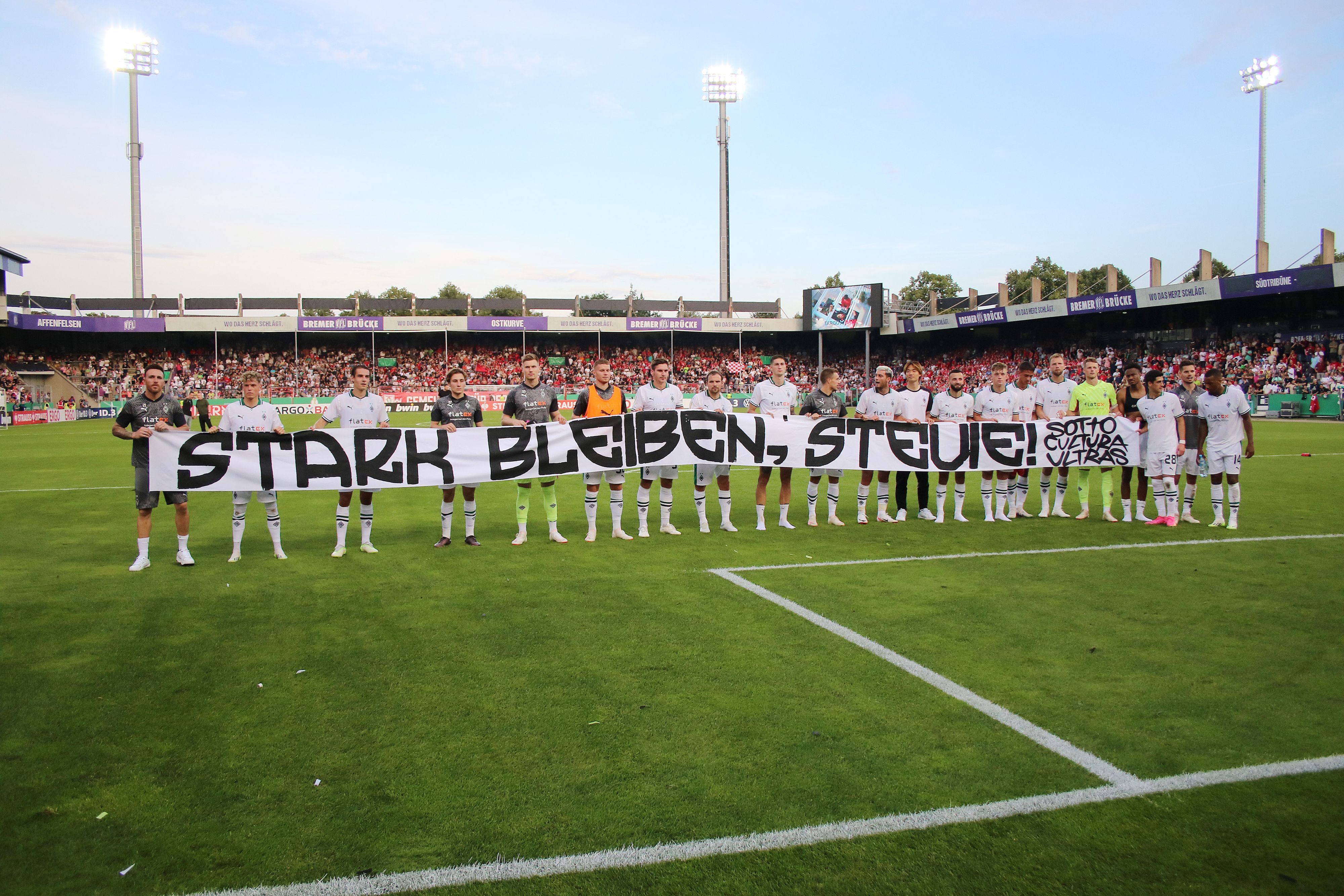 Die Mannschaft von Borussia Mönchengladbach hält ein Banner mit der Aufschrift „Stark bleiben, Stevie!“. Das Foto stammt vom 11. August 2023, im Anschluss an das DFB-Pokalspiel beim TuS Bersenbrück in Osnabrück.