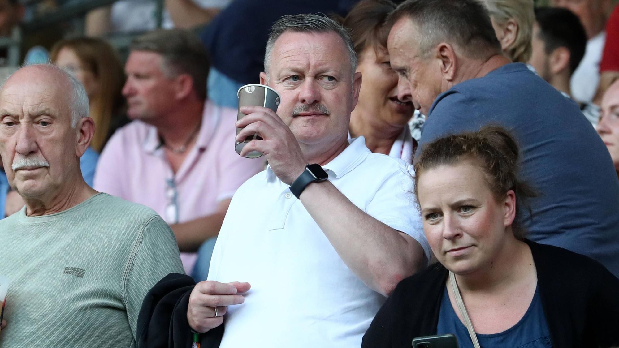 Roland Virkus hält am 12. August 2023 einen Kaffeebecher in der Hand und schaut sich das Spiel zwischen Viktoria Köln und Werder Bremen im DFB-Pokal an.