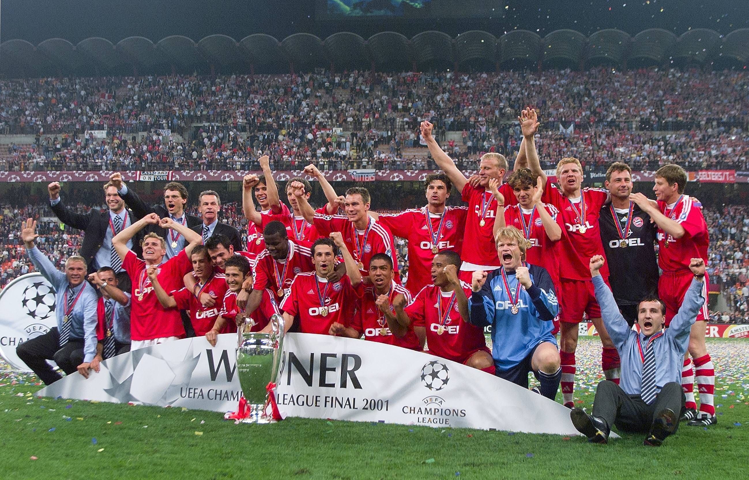Die Bayern-Spieler stehen hinter dem Champions League Pokal und jubeln. im Hintergrund sind die Tribünen des San Siro in Mailand zu sehen.