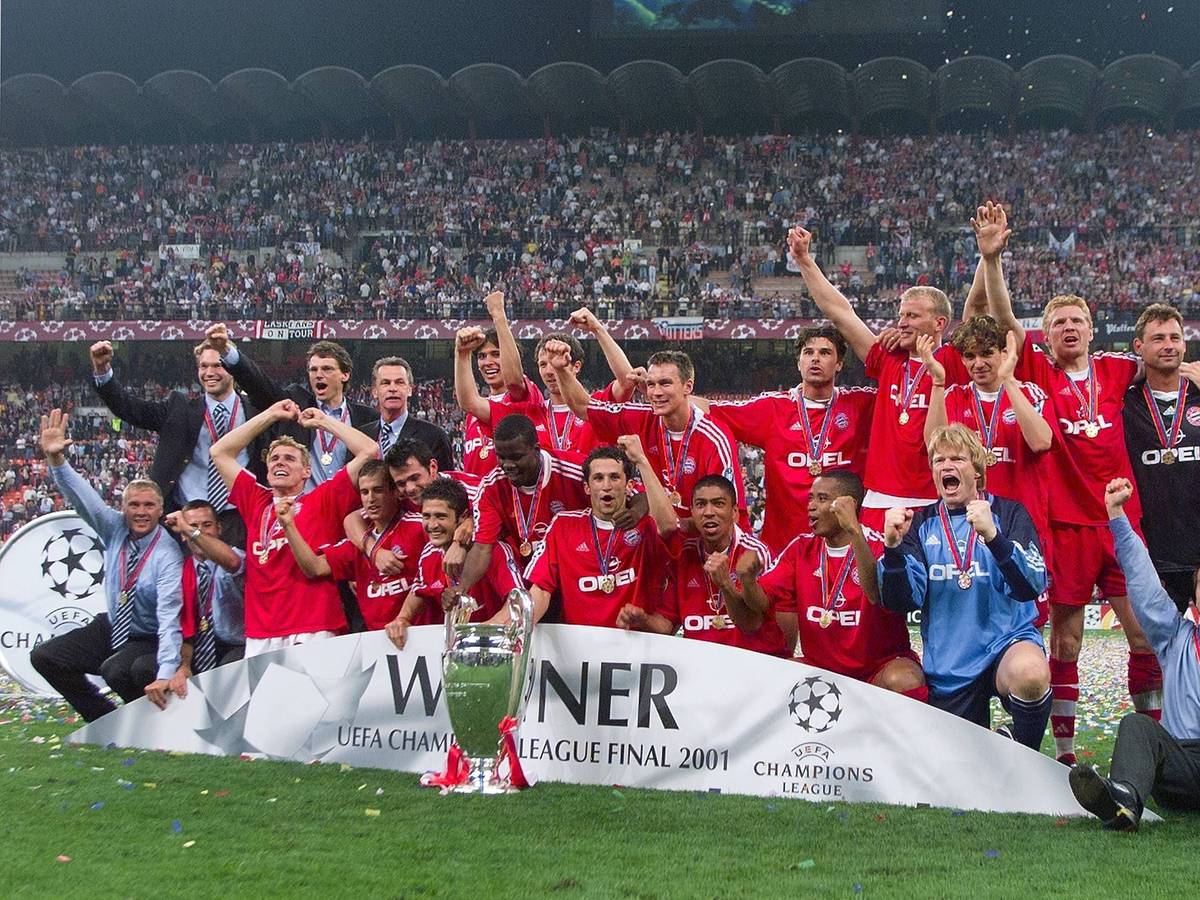 Die Bayern-Spieler stehen hinter dem Champions League Pokal und jubeln. im Hintergrund sind die Tribünen des San Siro in Mailand zu sehen.
