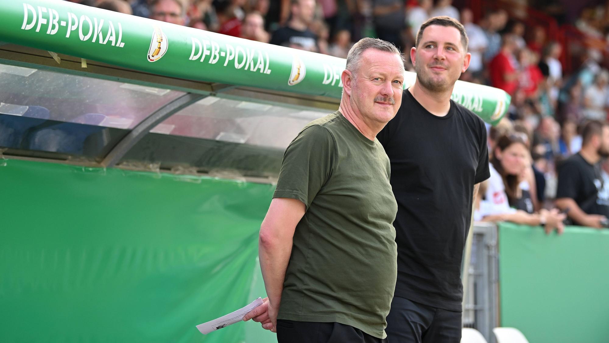 Roland Virkus und Nils Schmadtke stehen vor einer Ersatzbank.