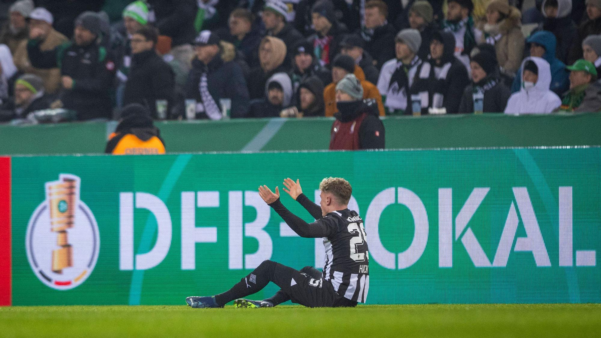 Robin Hack sitzt auf dem Spielfeld vor einer Bande mit der Aufschrift „DFB-Pokal“.