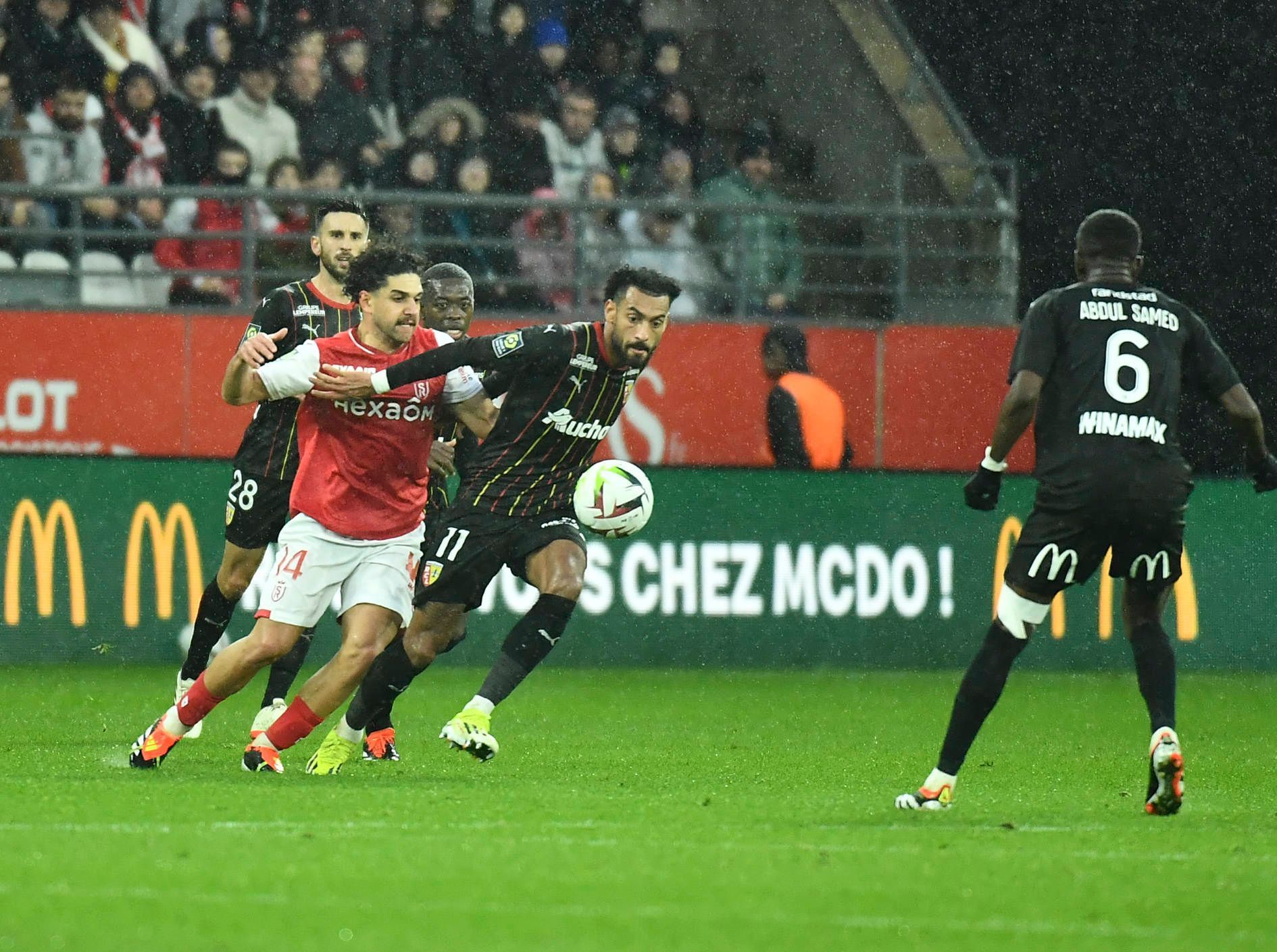 Reda Khadra im roten Reims-Trikot kämpft um den Ball mit Angelo Fulgini im schwarzen Lens-Trikot. Es regnet.