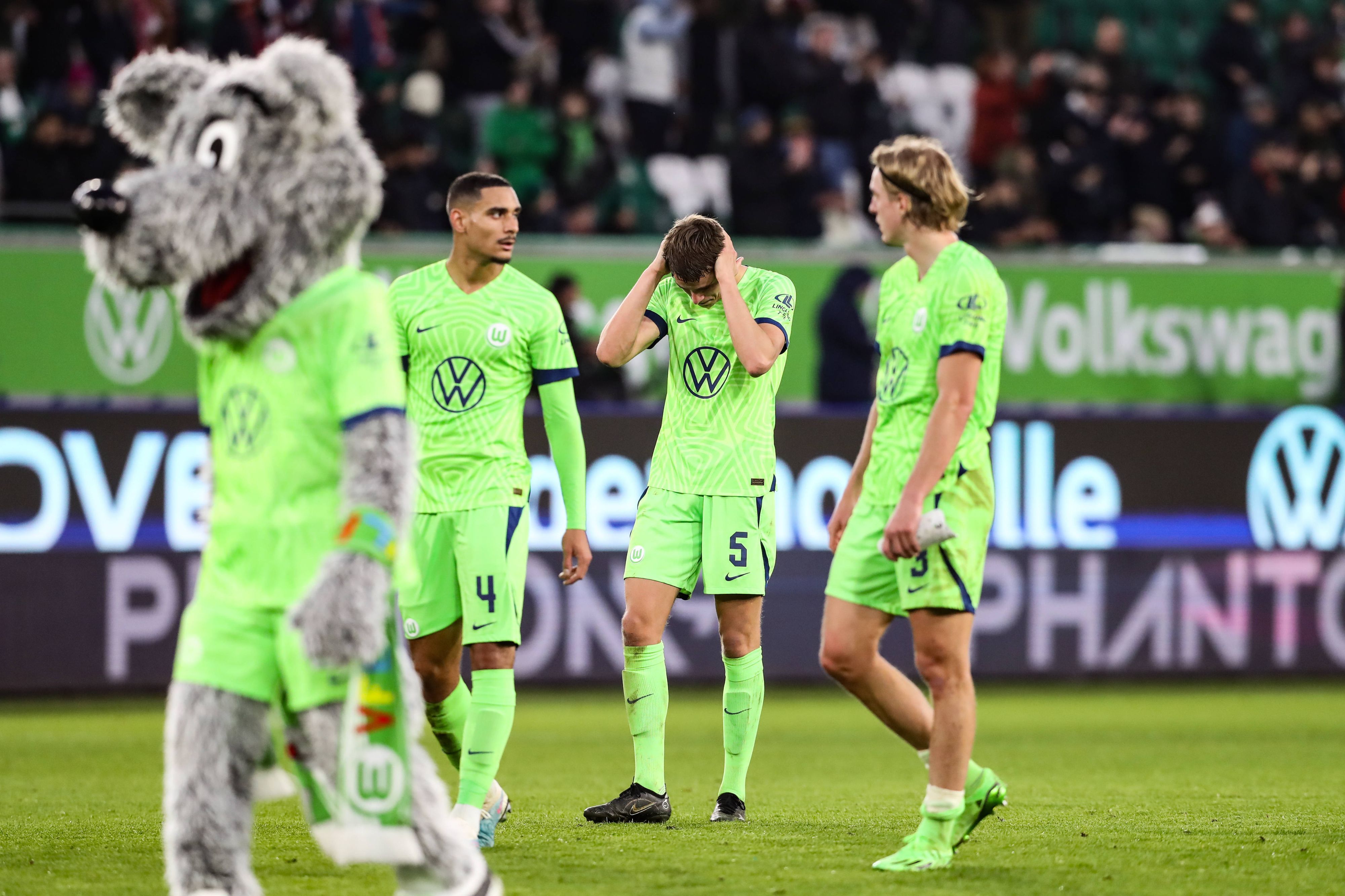 Drei Wolfsburg-Spieler ärgern sich. Auf der linken Seite läuft das Maskottchen Wölfi über den Platz.