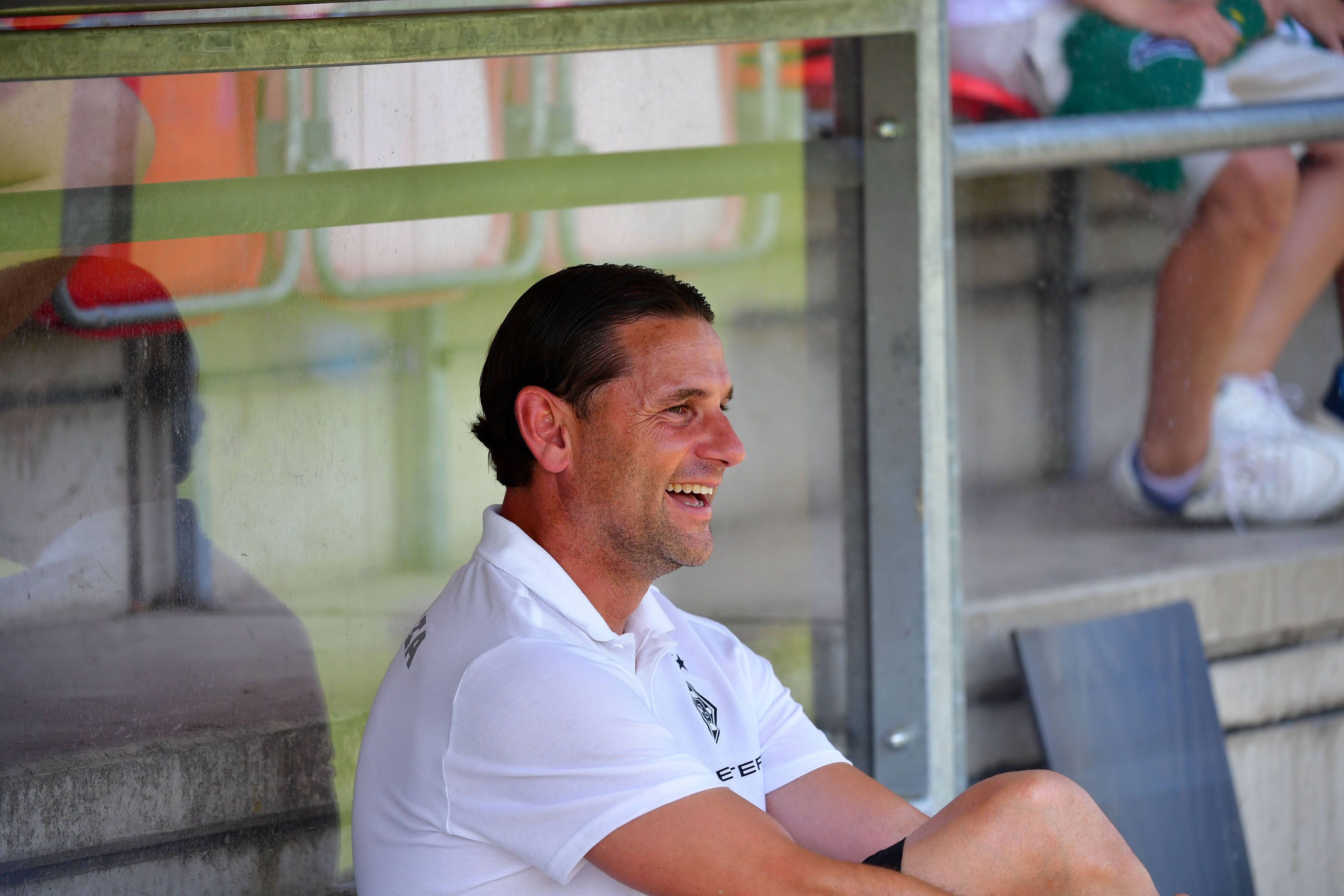 Gerardo Seoane sitzt auf der Trainerbank und lacht.