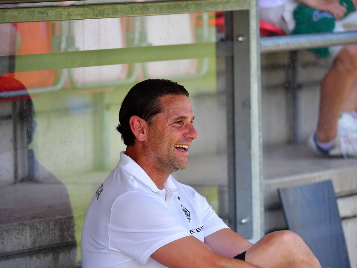Gerardo Seoane sitzt auf der Trainerbank und lacht.
