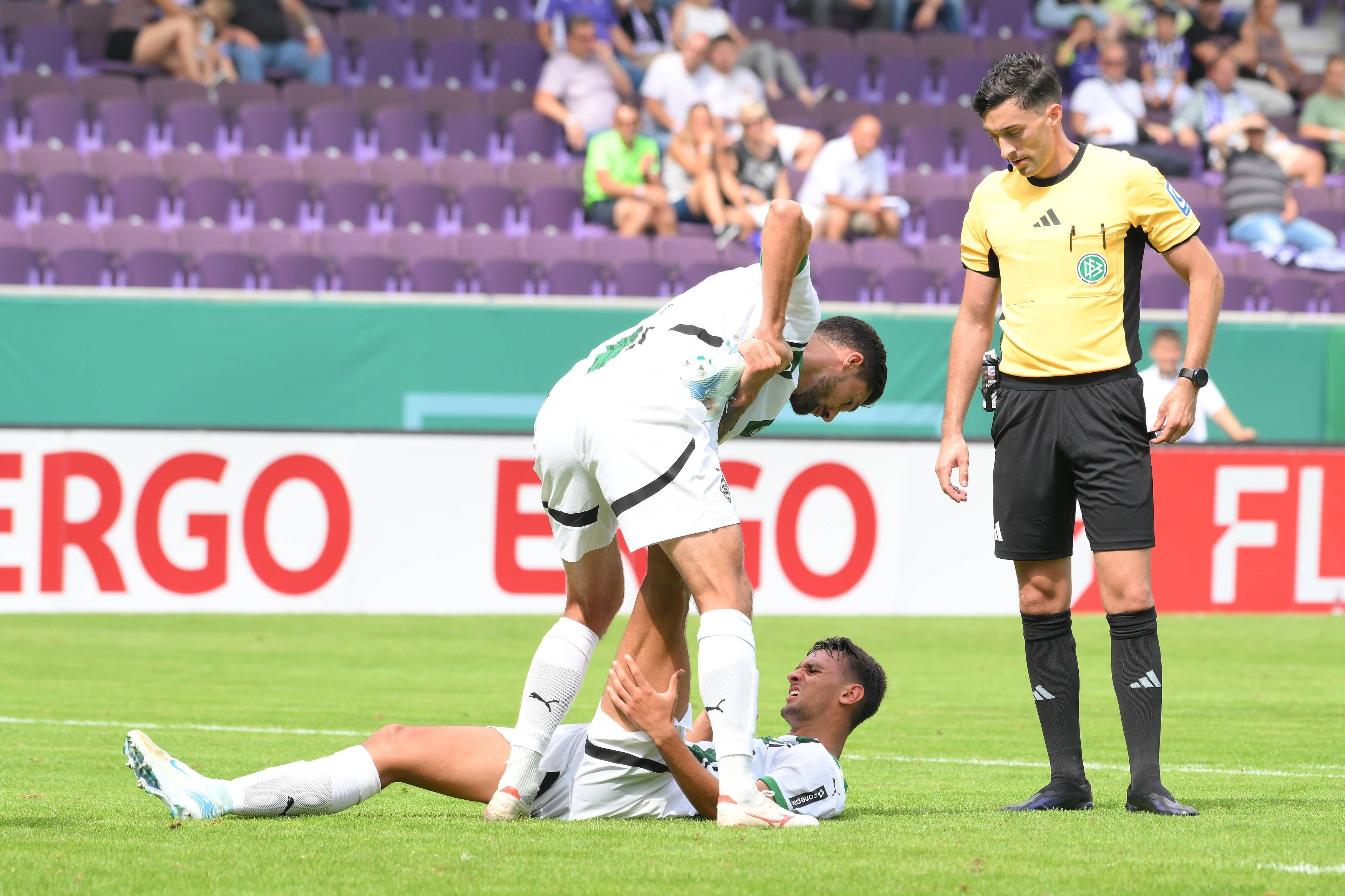 Borussia Mönchengladbachs Fabio Chiarodia liegt am Boden, Tim Kleindienst hilft ihm beim Dehnen. Schiedsrichter Florian Badstübner beobachtet die Szene.