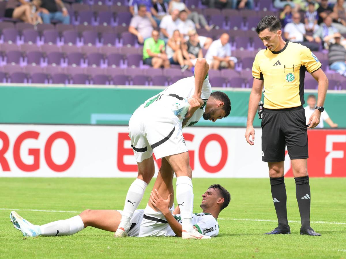 Borussia Mönchengladbachs Fabio Chiarodia liegt am Boden, Tim Kleindienst hilft ihm beim Dehnen. Schiedsrichter Florian Badstübner beobachtet die Szene.