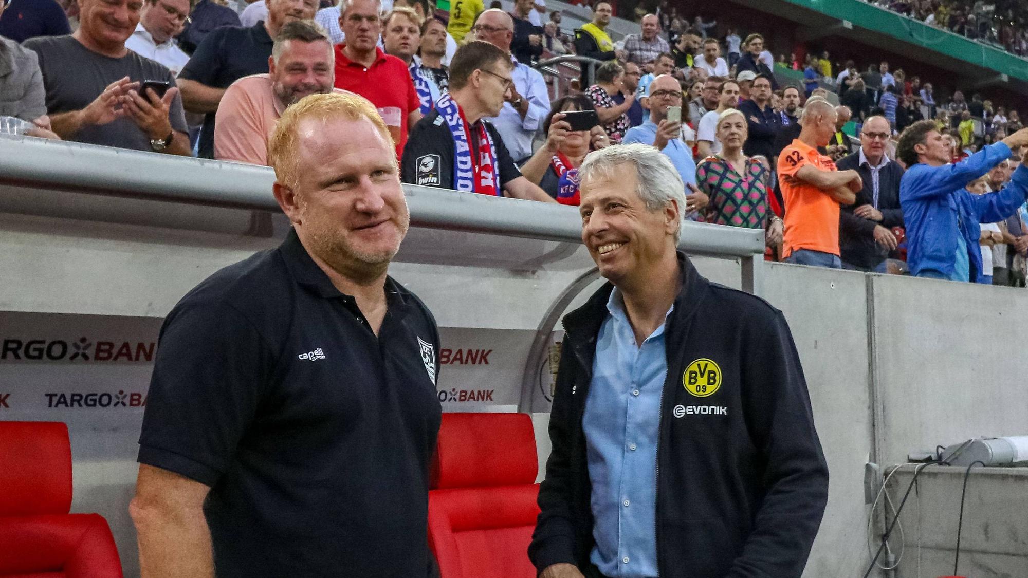 Uerdingens Trainer Heiko Vogel und Dortmunds Trainer Lucien Favre stehen vor dem Pokalspiel lachend nebeneinander an der Trainerbank.