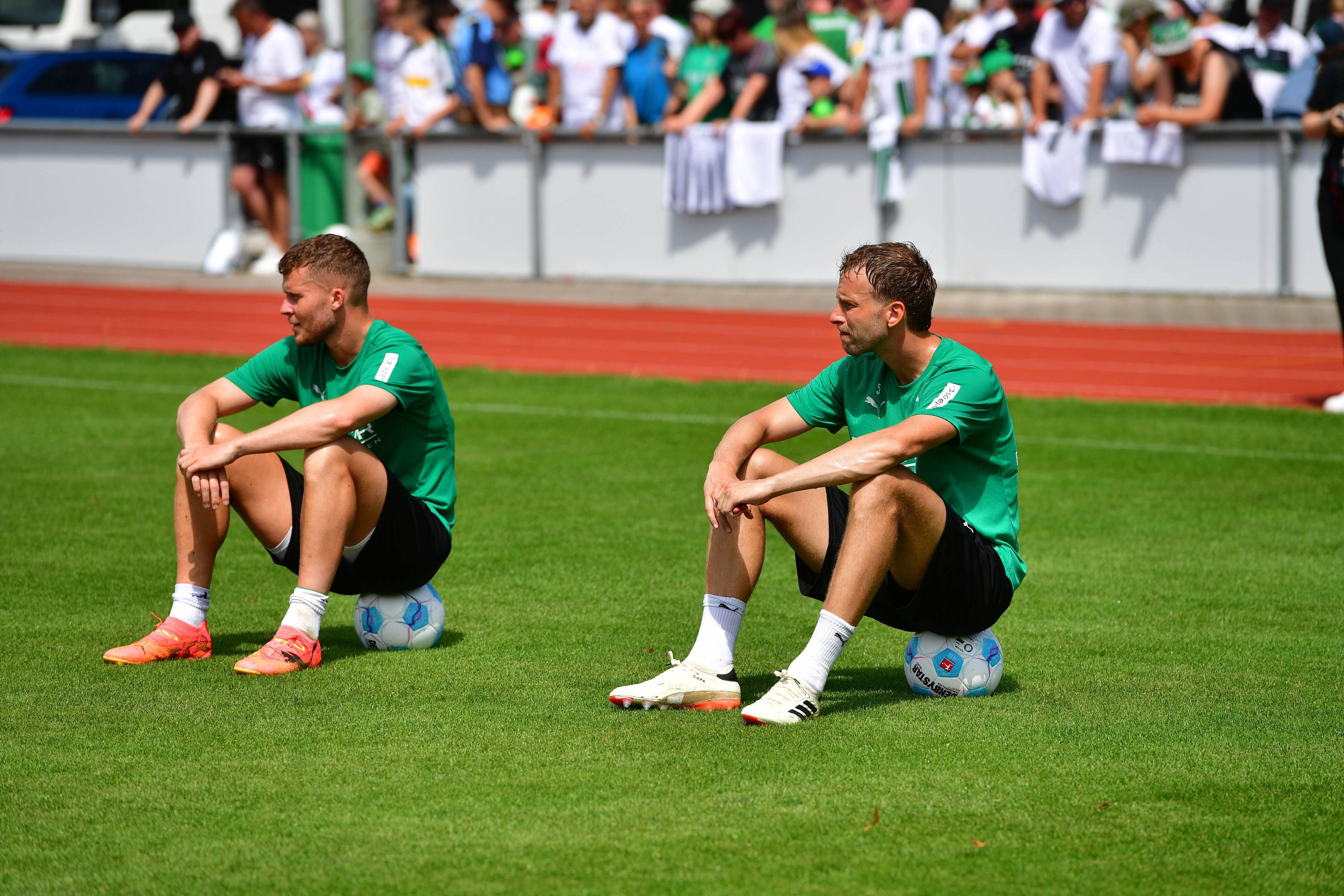 Die Gladbach-Profis Nico Elvedi und Marvin Friedrich und sitzen auf dem Trainingsplatz nebeneinander auf Fußbällen.