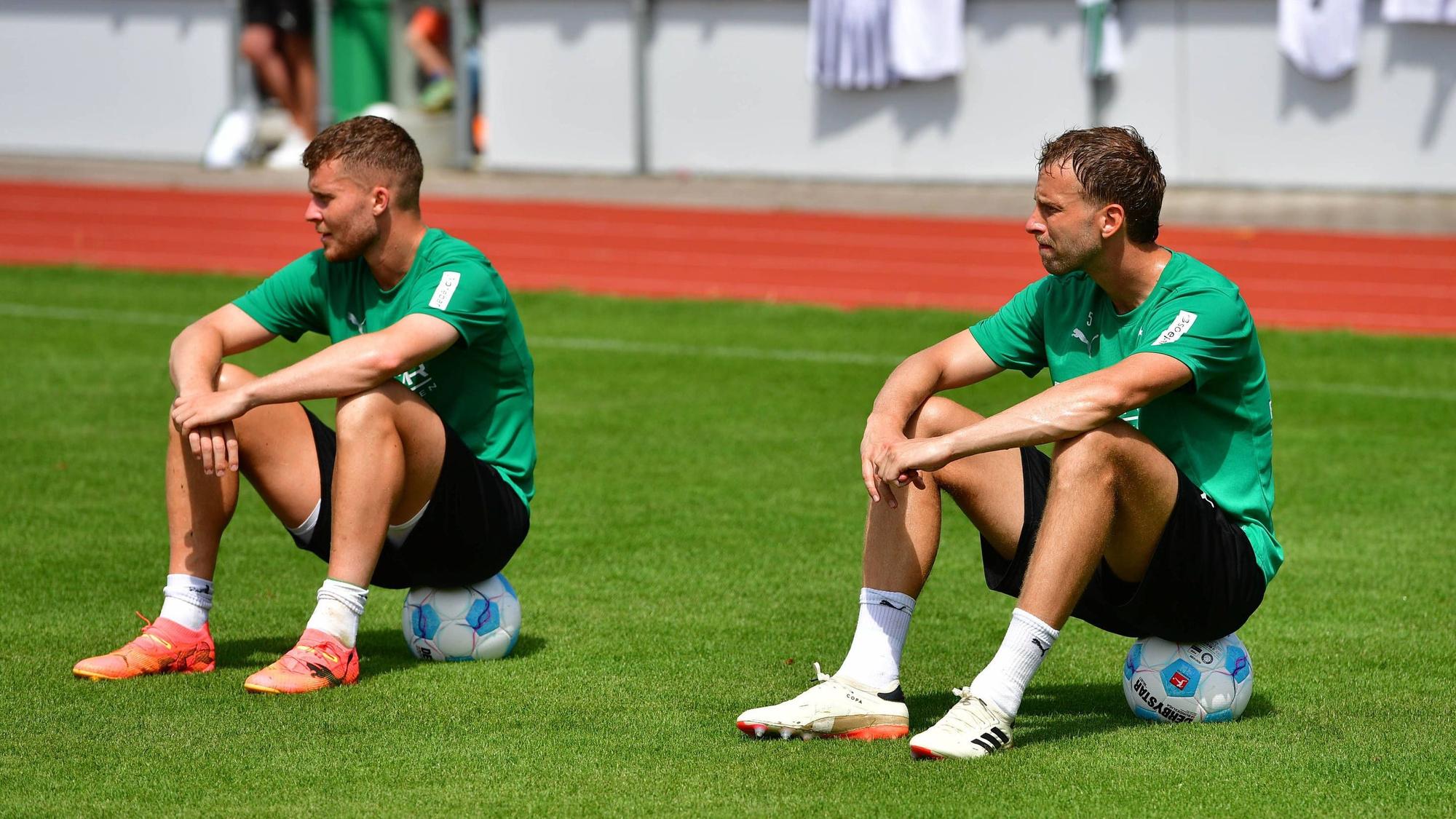 Die Gladbach-Profis Nico Elvedi und Marvin Friedrich und sitzen auf dem Trainingsplatz nebeneinander auf Fußbällen.