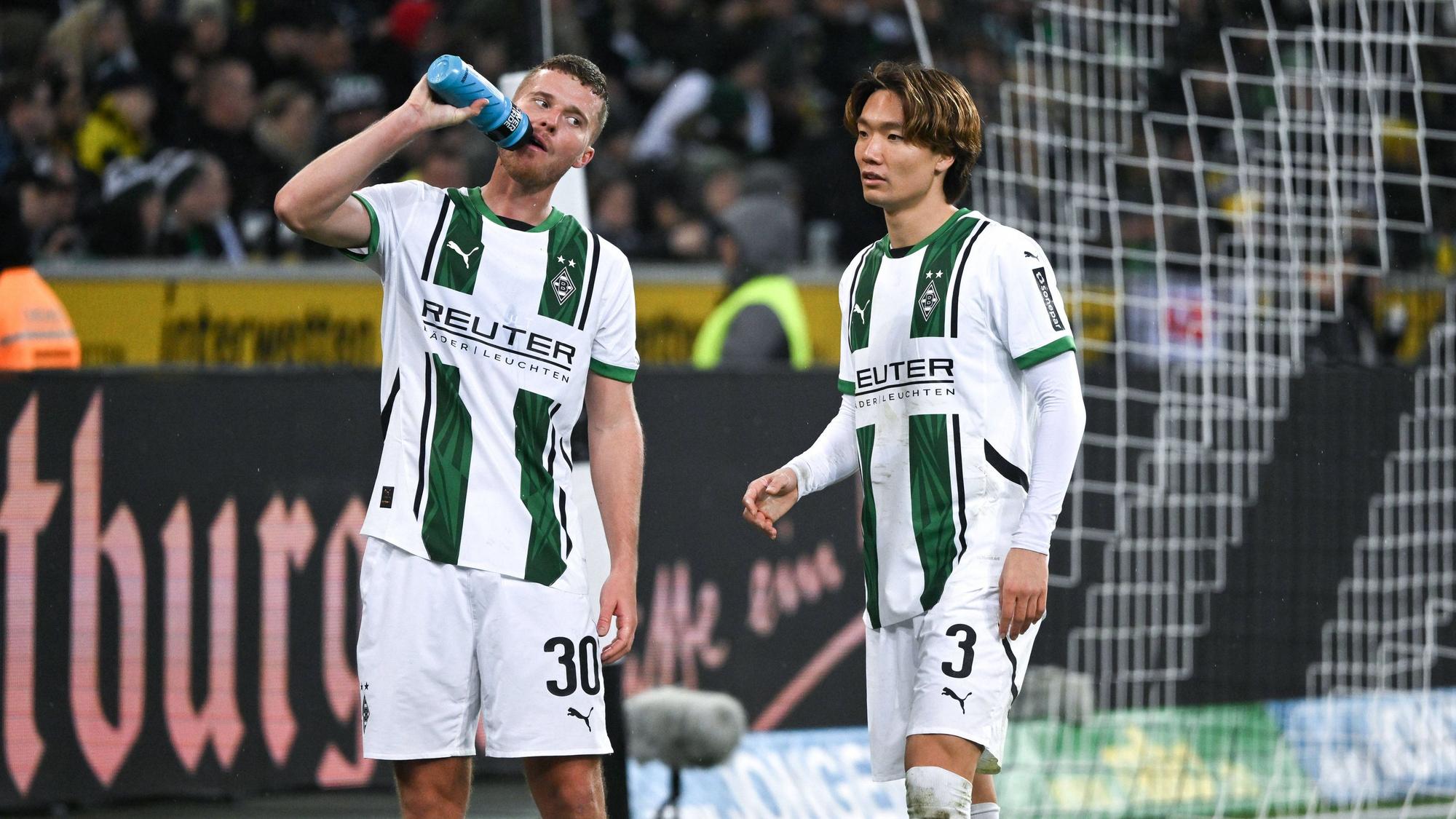 Die Gladbach-Profis Nico Elvedi und Ko Itakura stehen beim Spiel gegen Borussia Dortmund nebeneinander, Elvedi trinkt aus einer Wasserflasche.