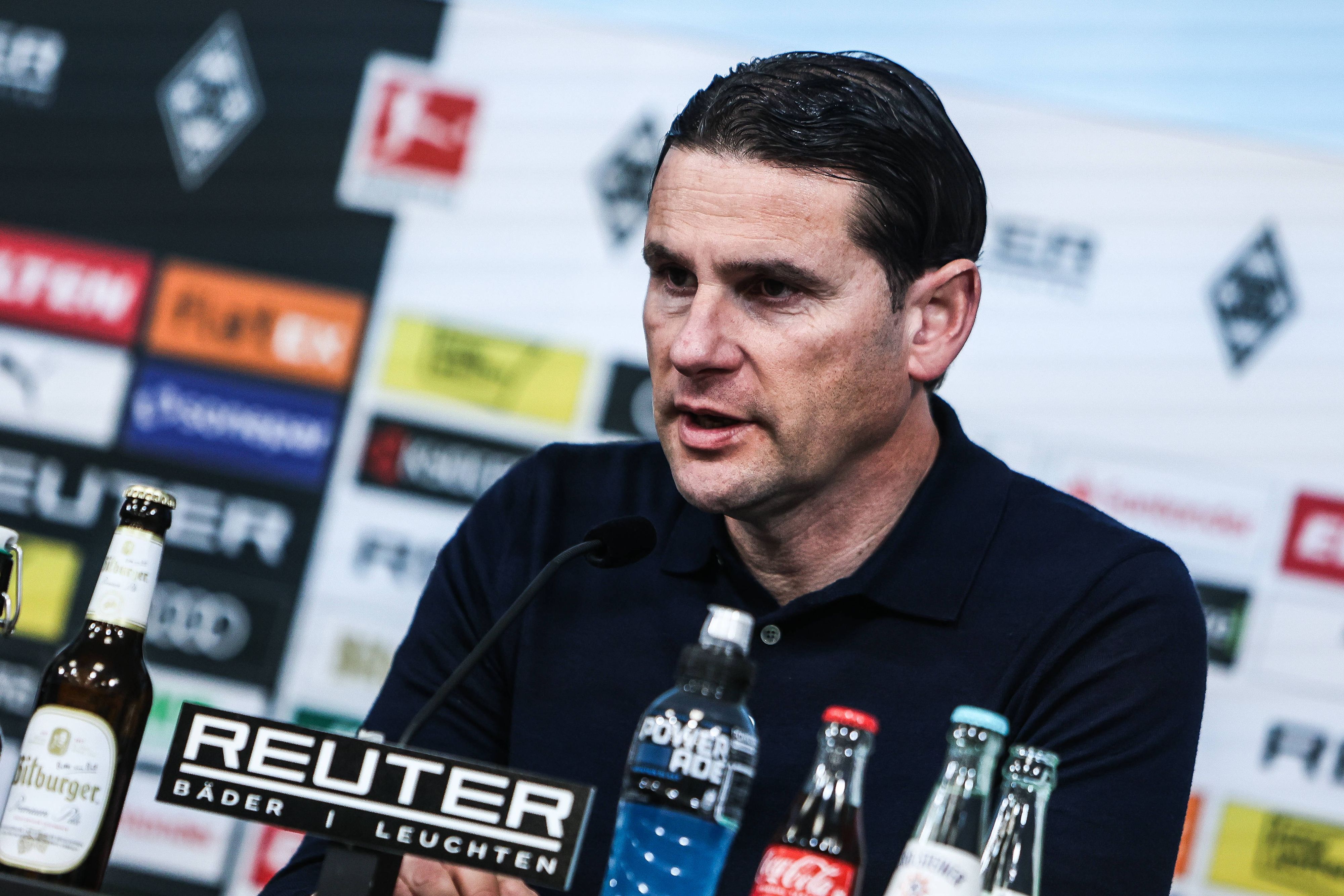 Borussia Mönchengladbachs Trainer sitzt bei der Pressekonferenz auf dem Podium und spricht ins Mikrofon.