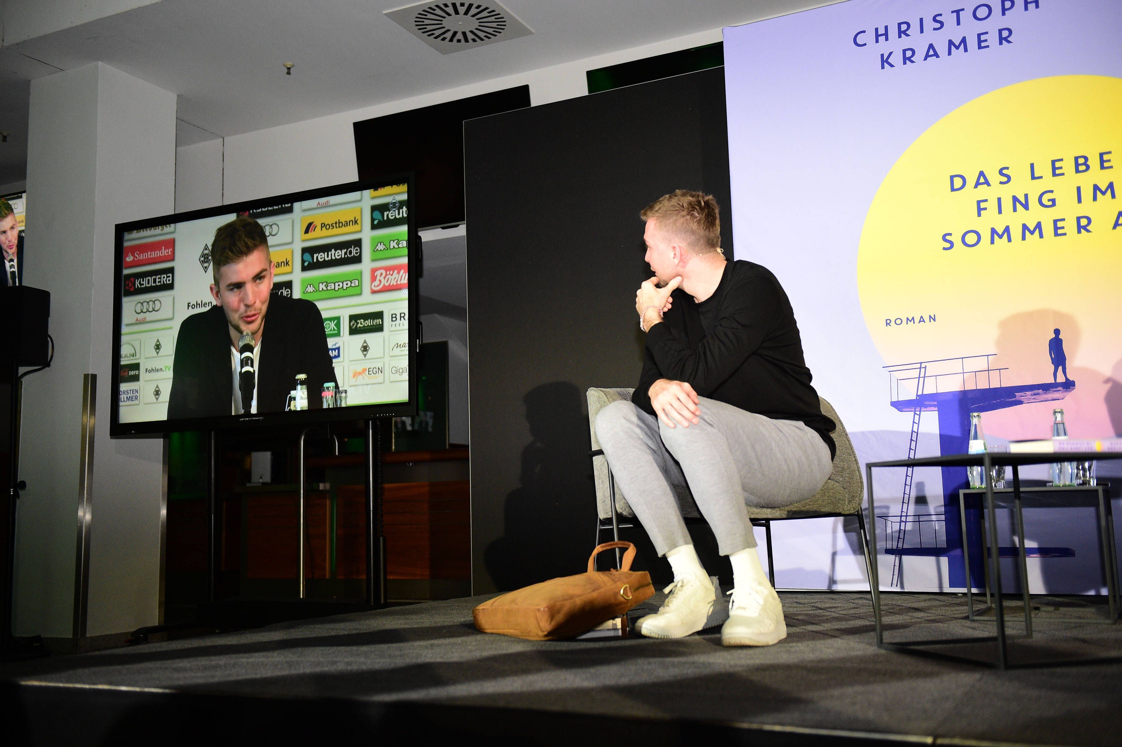Christoph Kramer sitzt bei einer Lesung auf der Bühne, ihm Hintergrund werden auf einem Bildschirm Szenen seiner Karriere gezeigt.
