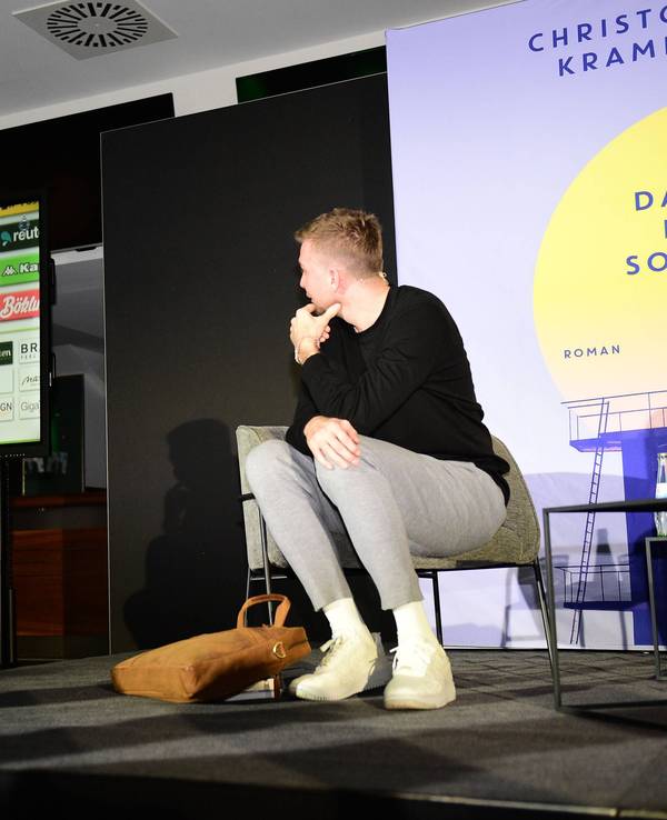 Christoph Kramer sitzt bei einer Lesung auf der Bühne, ihm Hintergrund werden auf einem Bildschirm Szenen seiner Karriere gezeigt.