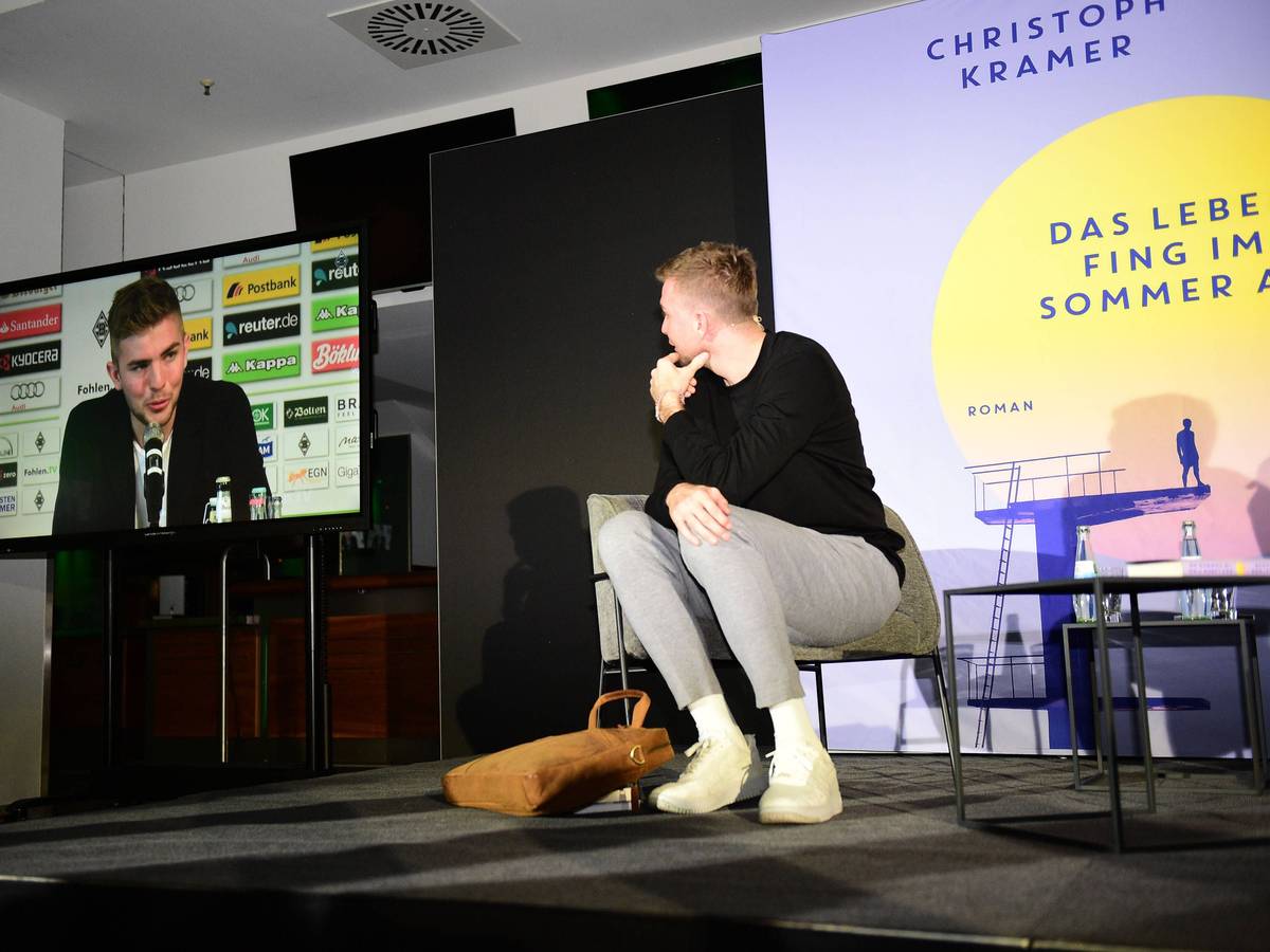 Christoph Kramer sitzt bei einer Lesung auf der Bühne, ihm Hintergrund werden auf einem Bildschirm Szenen seiner Karriere gezeigt.