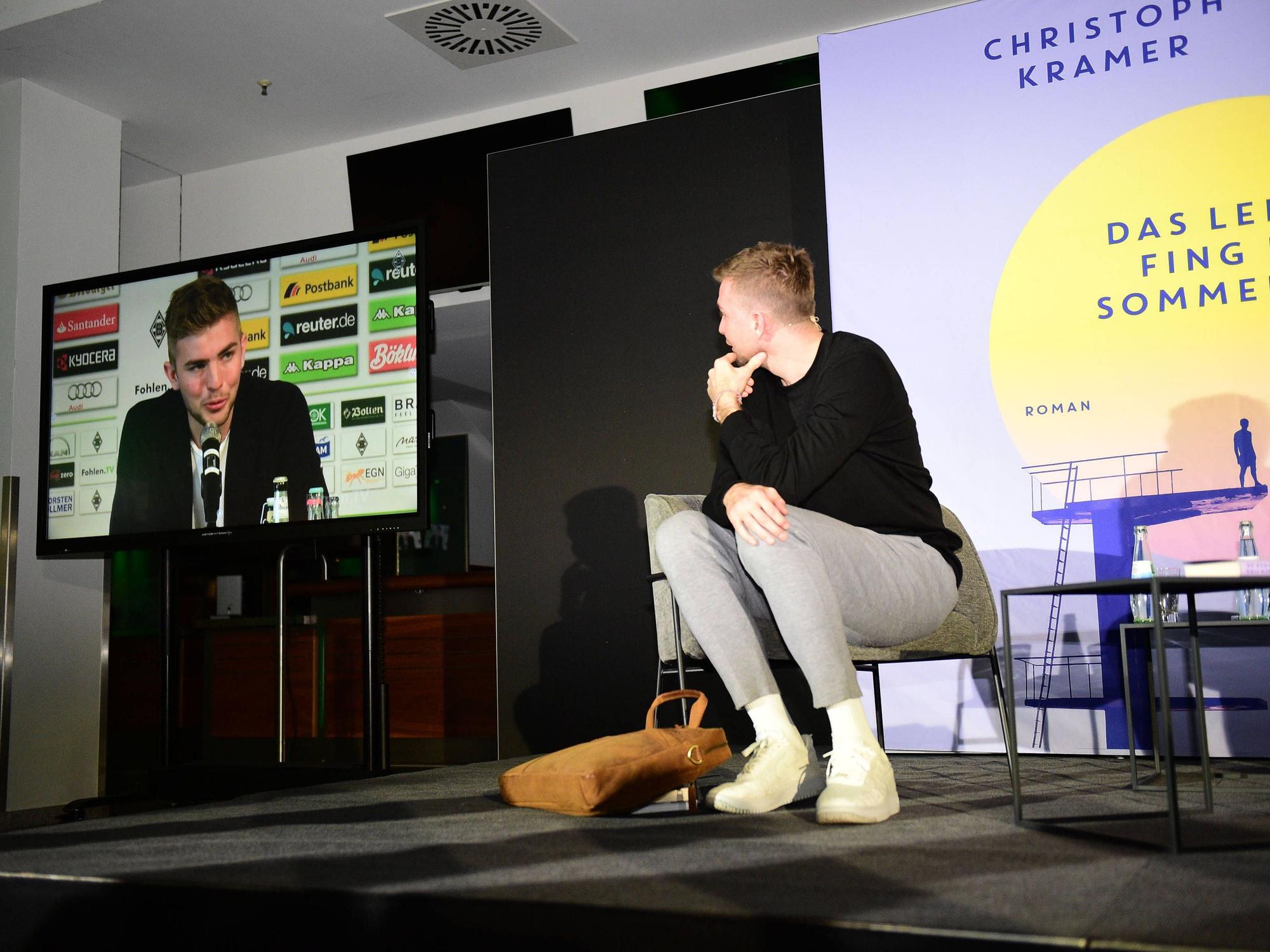 Christoph Kramer sitzt bei einer Lesung auf der Bühne, ihm Hintergrund werden auf einem Bildschirm Szenen seiner Karriere gezeigt.