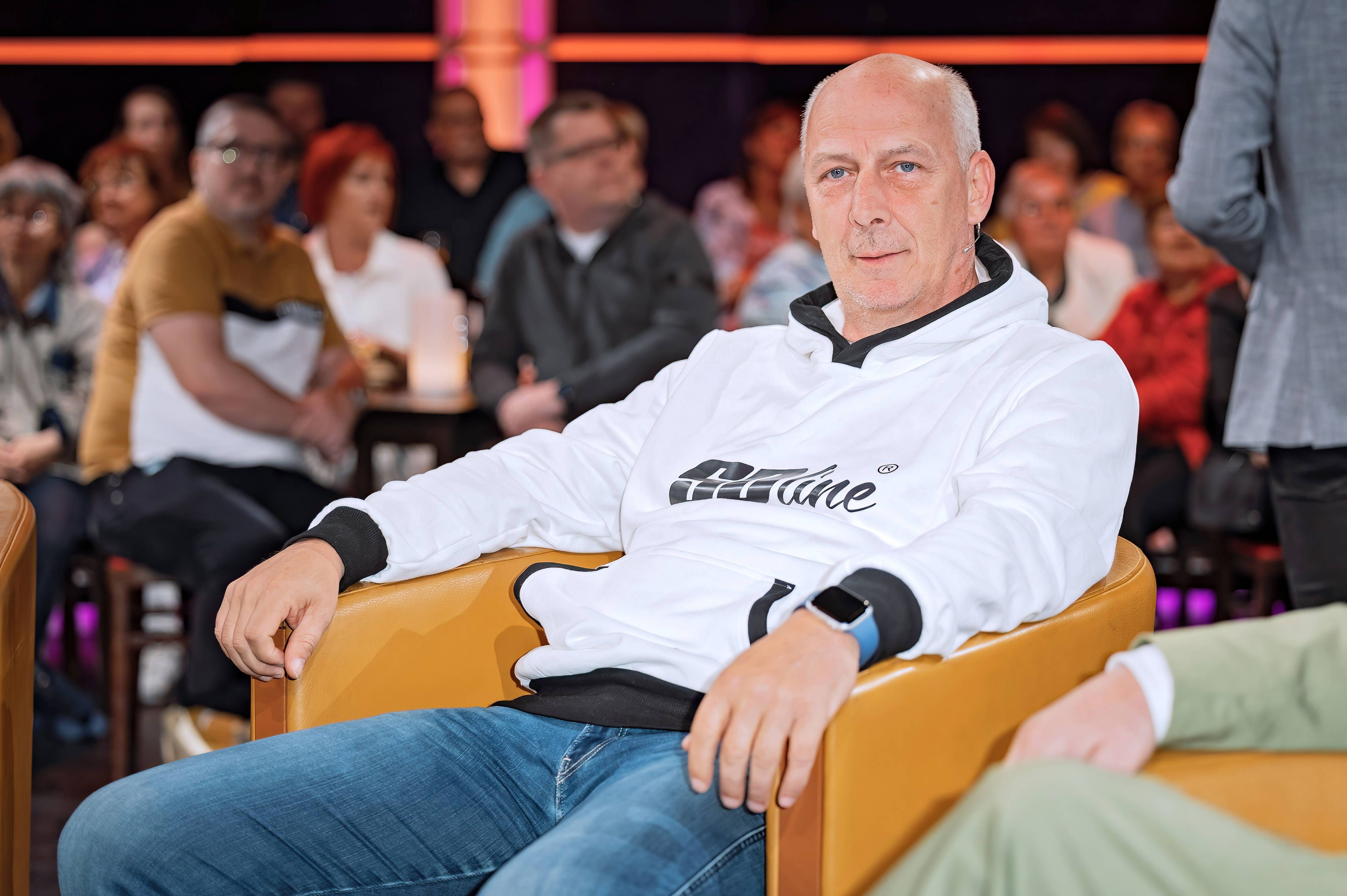 Mario Basler sitzt auf einem senffarbenen Sessel und hat beide Arme auf den Armlehnen liegen.