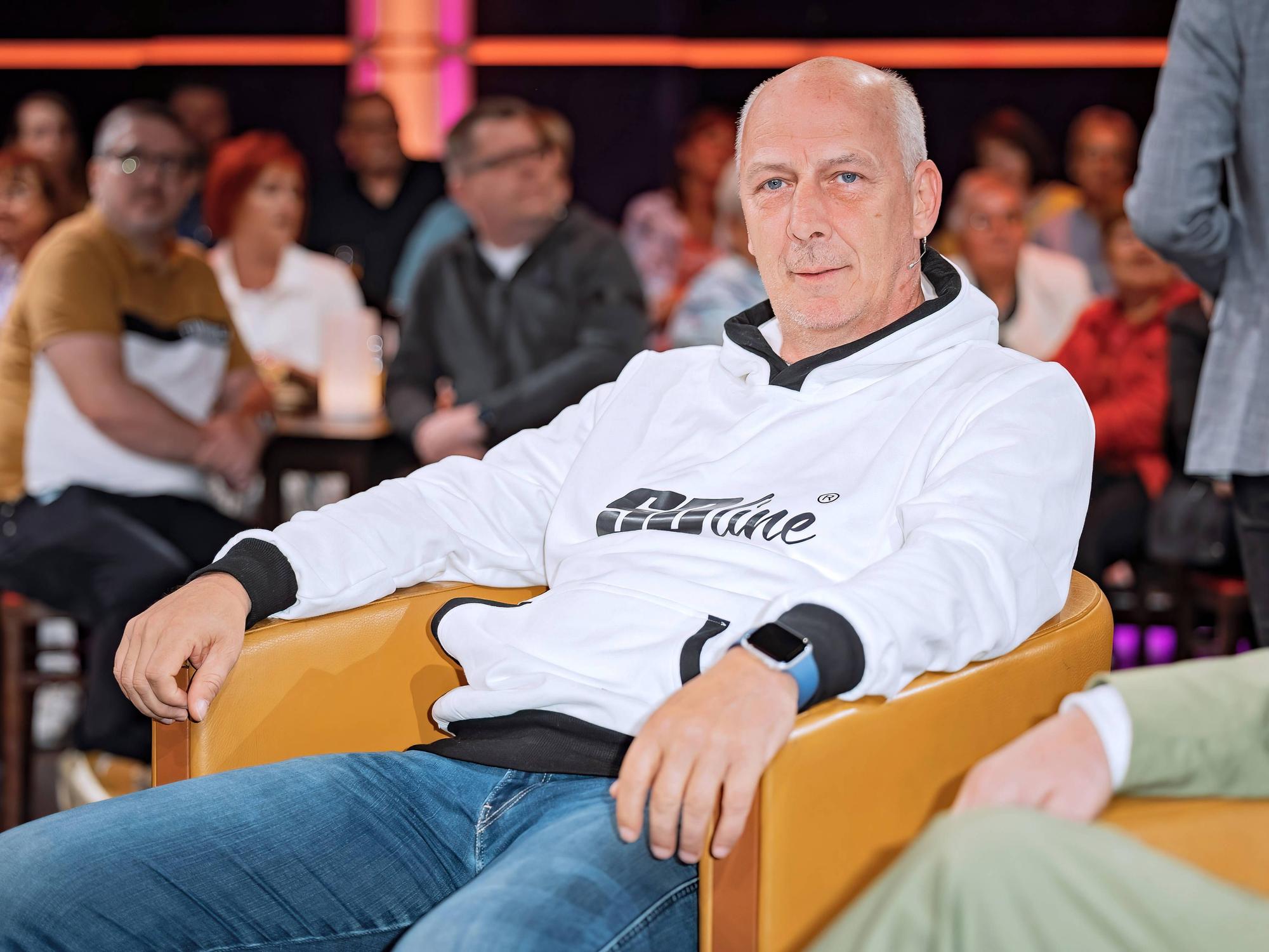 Mario Basler sitzt auf einem senffarbenen Sessel und hat beide Arme auf den Armlehnen liegen.