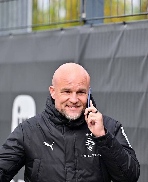 Rouven Schröder telefoniert in Gladbach-Jacke am Trainingsplatz und grinst dabei.