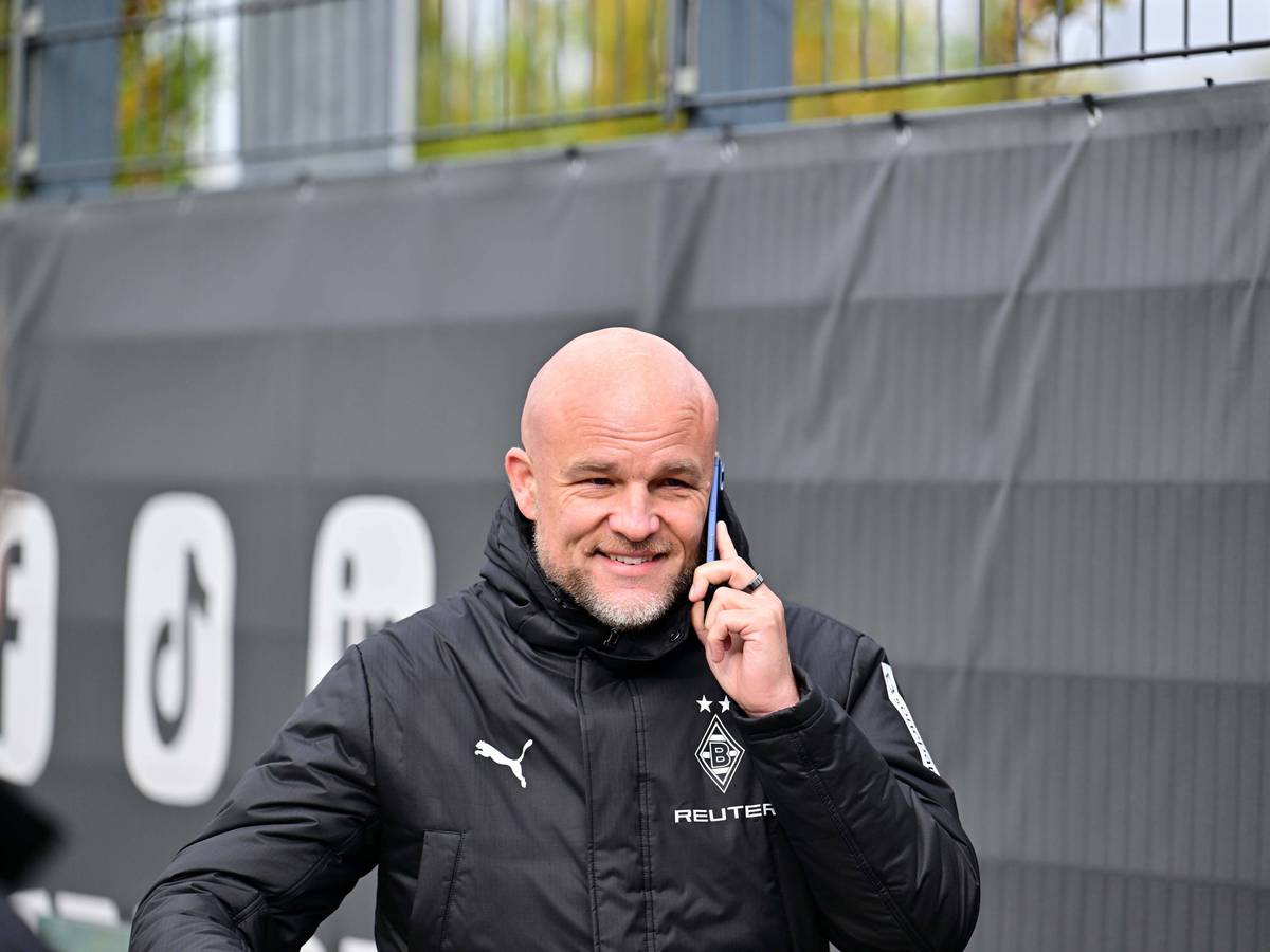 Rouven Schröder telefoniert in Gladbach-Jacke am Trainingsplatz und grinst dabei.